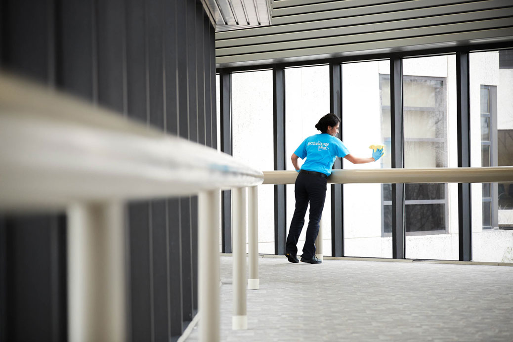 Two cleaning people are cleaning the inside of a classroom by wiping down desks and using a microfiber mop to wipe dust and debris off floors
